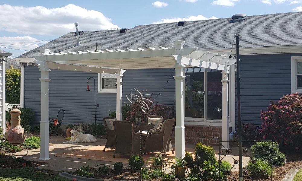 White pergola over patio with wicker chairs and a dog, surrounded by garden plants and a blue house exterior.