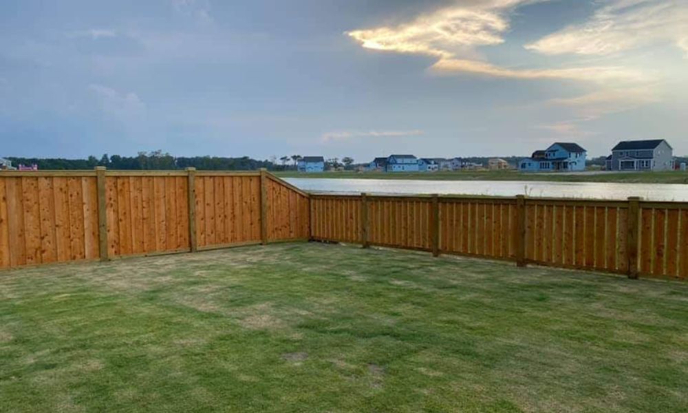 Fenced backyard with green lawn, view of a lake, and houses under a cloudy sky. Peaceful and scenic landscape.