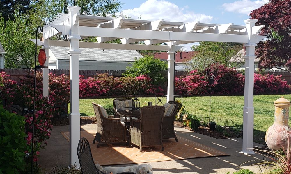 White pergola in a garden setting with outdoor dining and a dog relaxing. Bright flowers and plants surround the area.