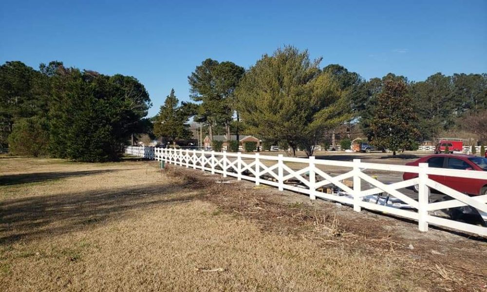 White fence along a grassy field with trees and parked cars in the background under a clear blue sky.